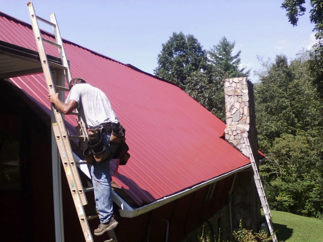 Roofer on a ladder beside a red metal roof and stone chimney — Soffit & Fascia Repair by Roofing Brothers in Indian Mound